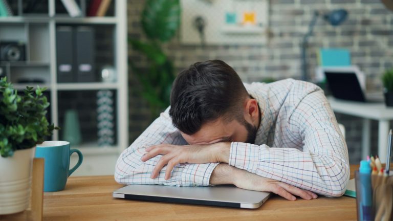 "Seorang pria tampak kelelahan dan tertidur di atas laptop di meja kerja, menunjukkan rasa lelah atau kurang motivasi saat bekerja di kantor."